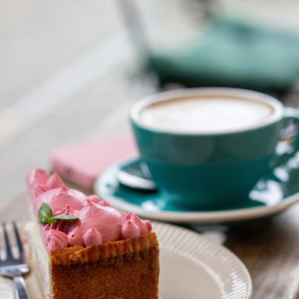 Home A slice of cake with pink frosting and a cappuccino in a blue cup on a wooden table.